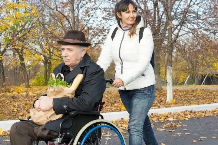 Woman wearing a white jacket pushing a senior man in a wheelchair with his grocery items on his lap